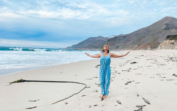 Paula Wild on the beach with arms up sharing information about lessons learning in online yoga teacher training