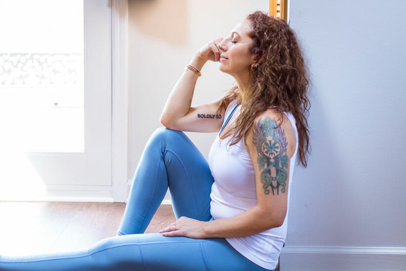 Kate Evans sitting with hand on head wearing white tank top and blue yoga pants