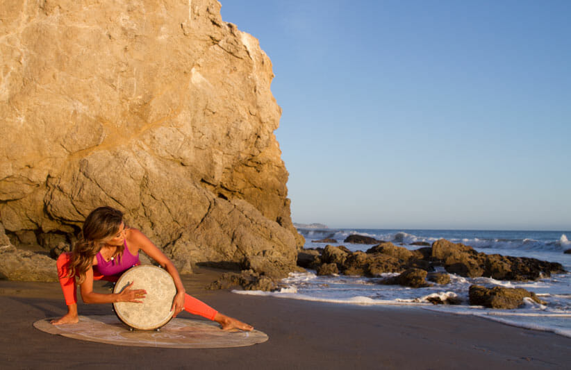 Desi Barlett playing a hand drum by the ocean wearing red yoga clothes in front of rocky coastline