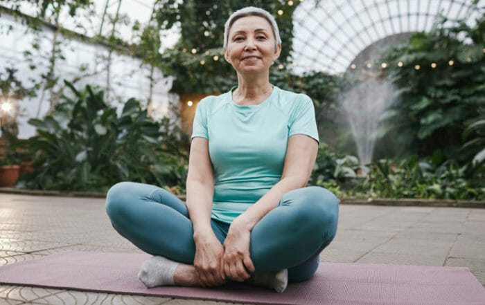 Woman smiling on yoga mat wearing yoga clothes around plants demonstrating healthy yoga for degenerative disc disease