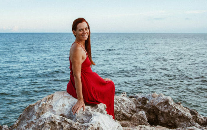 Enicia Fisher wearing red dress sitting on rocks by the ocean demonstrating the wellness benefits of the beach