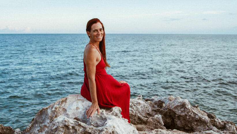 Enicia Fisher wearing red dress sitting on rocks by the ocean demonstrating the wellness benefits of the beach