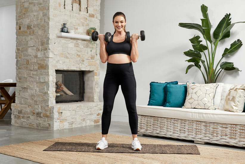 Emily Skye wearing black yoga clothes lifting dumbbells while pregnant in living room