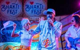 Bhakti Fest Kirtan Performers on stage. Man with white baseball cap holding microphone and woman dancing in front of Bhakti Fest stage signs.