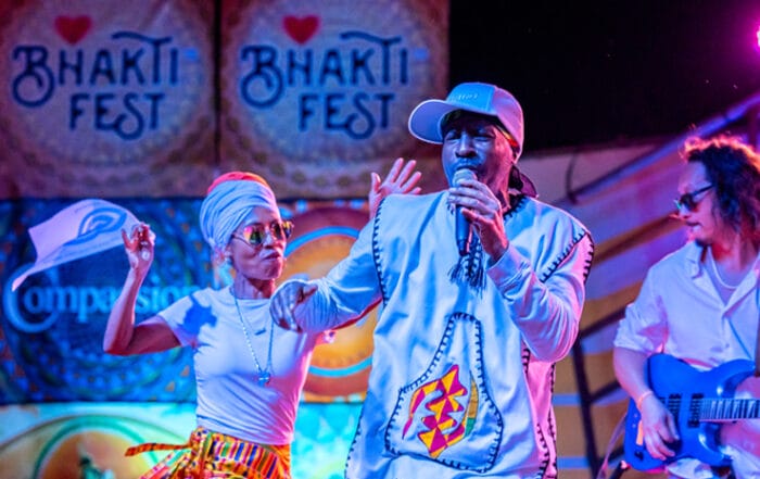 Bhakti Fest Kirtan Performers on stage. Man with white baseball cap holding microphone and woman dancing in front of Bhakti Fest stage signs.