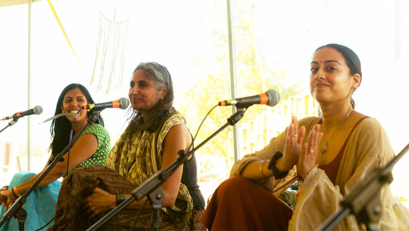 Three women singing with microphones sitting on a stage at Bhakti Fest