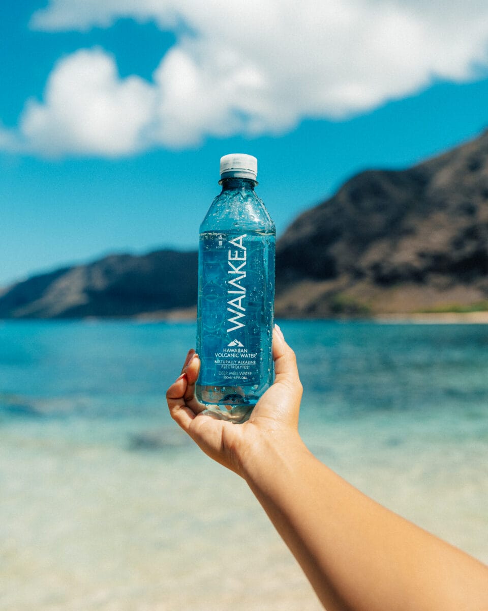 person holding Waiakea water bottle above a view of the beach